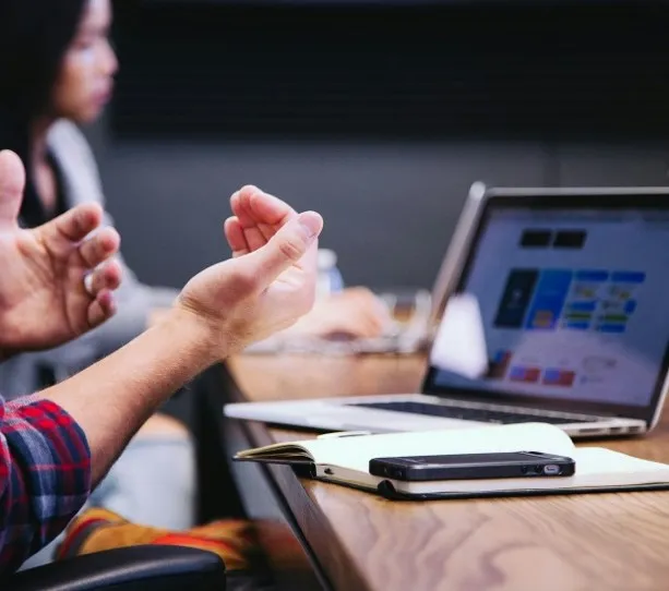 Iemand gebaart met de handen tijdens een overleg aan tafel met een laptop en een telefoon