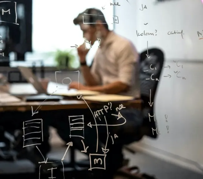 Schetsen van een systeemontwerp op een glazen wand, met daarachter iemand aan een bureau