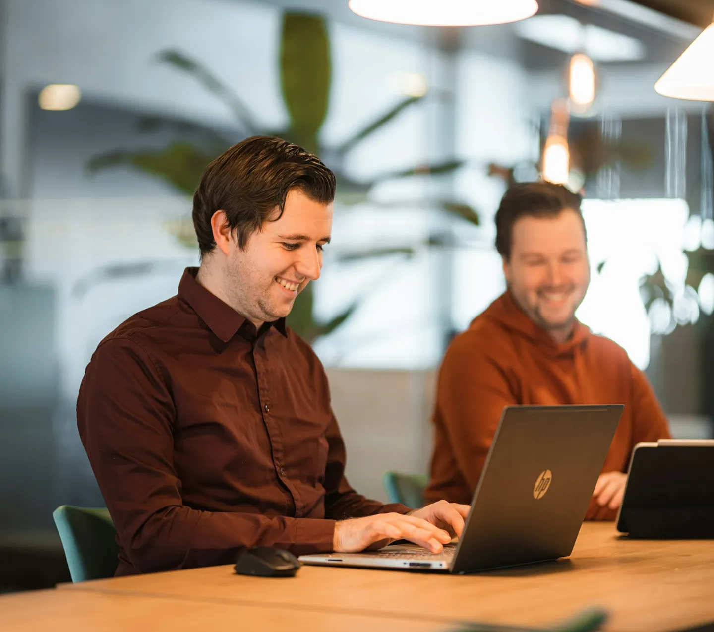 Twee lachende mannen aan een tafel, een van hen typt op een laptop