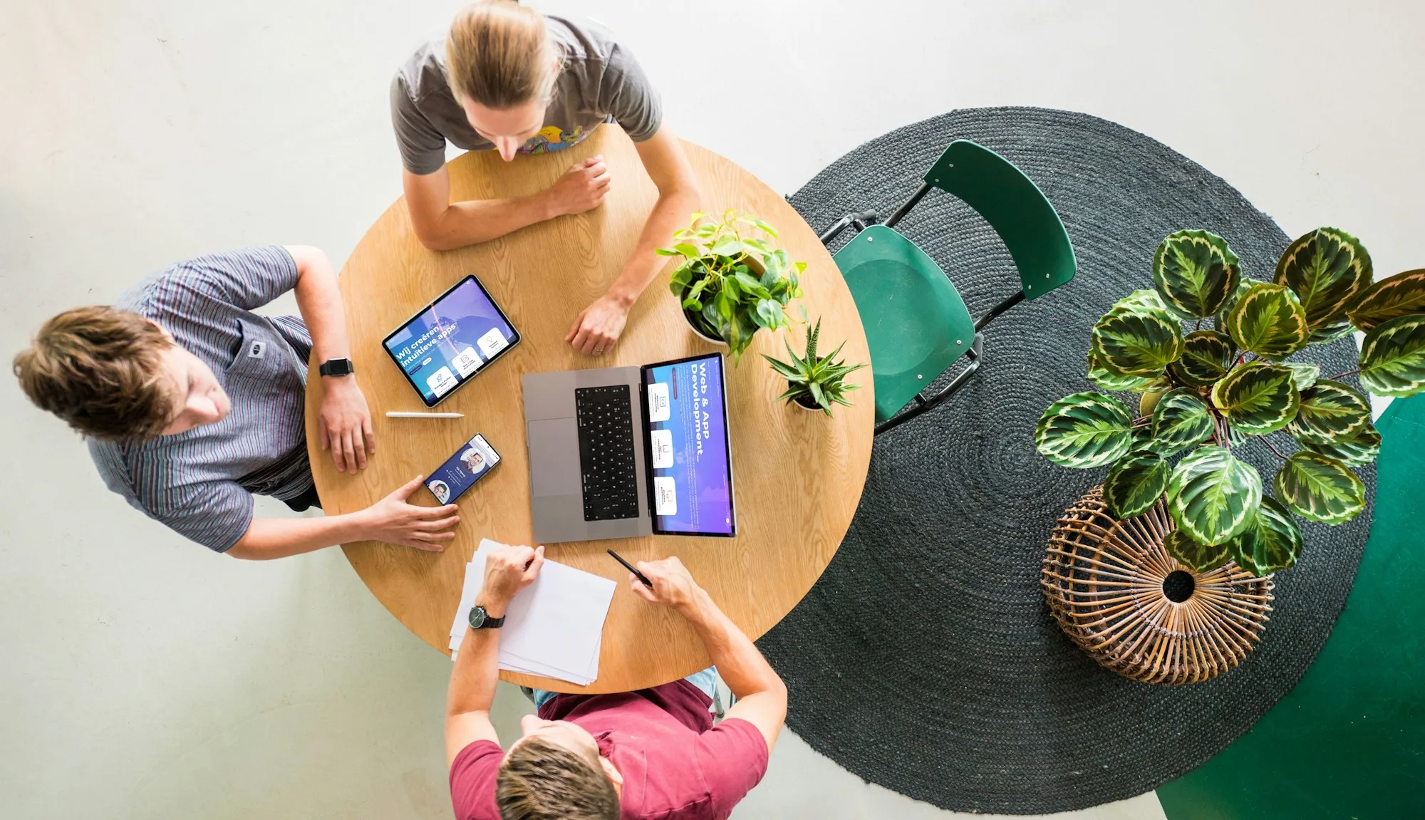 Three people talking at a round table with a laptop, tablet and phone, seen from above