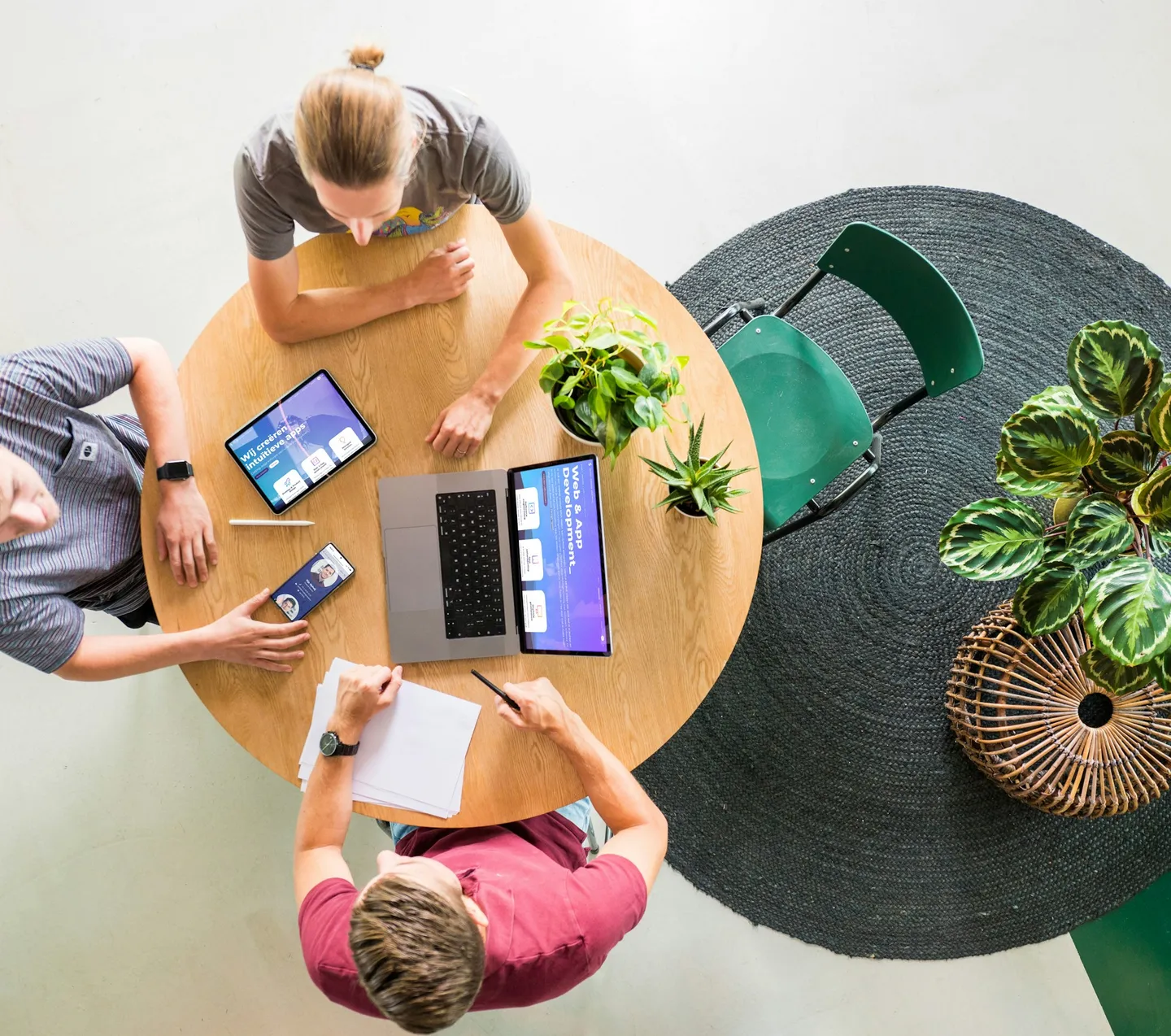 Three people talking at a round table with a laptop, tablet and phone, seen from above