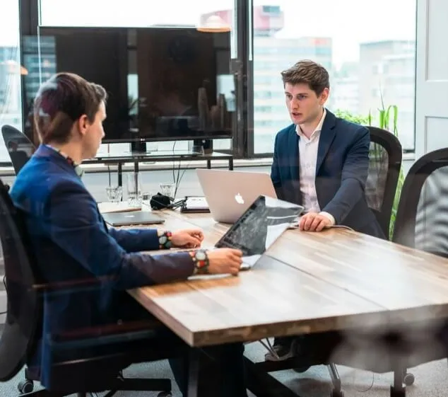Twee mannen in pak overleggen aan een tafel met laptops