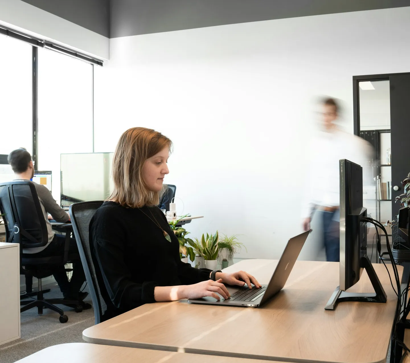 Een vrouw werkt op een laptop aan een bureau in een licht kantoor, met collega's op de achtergrond