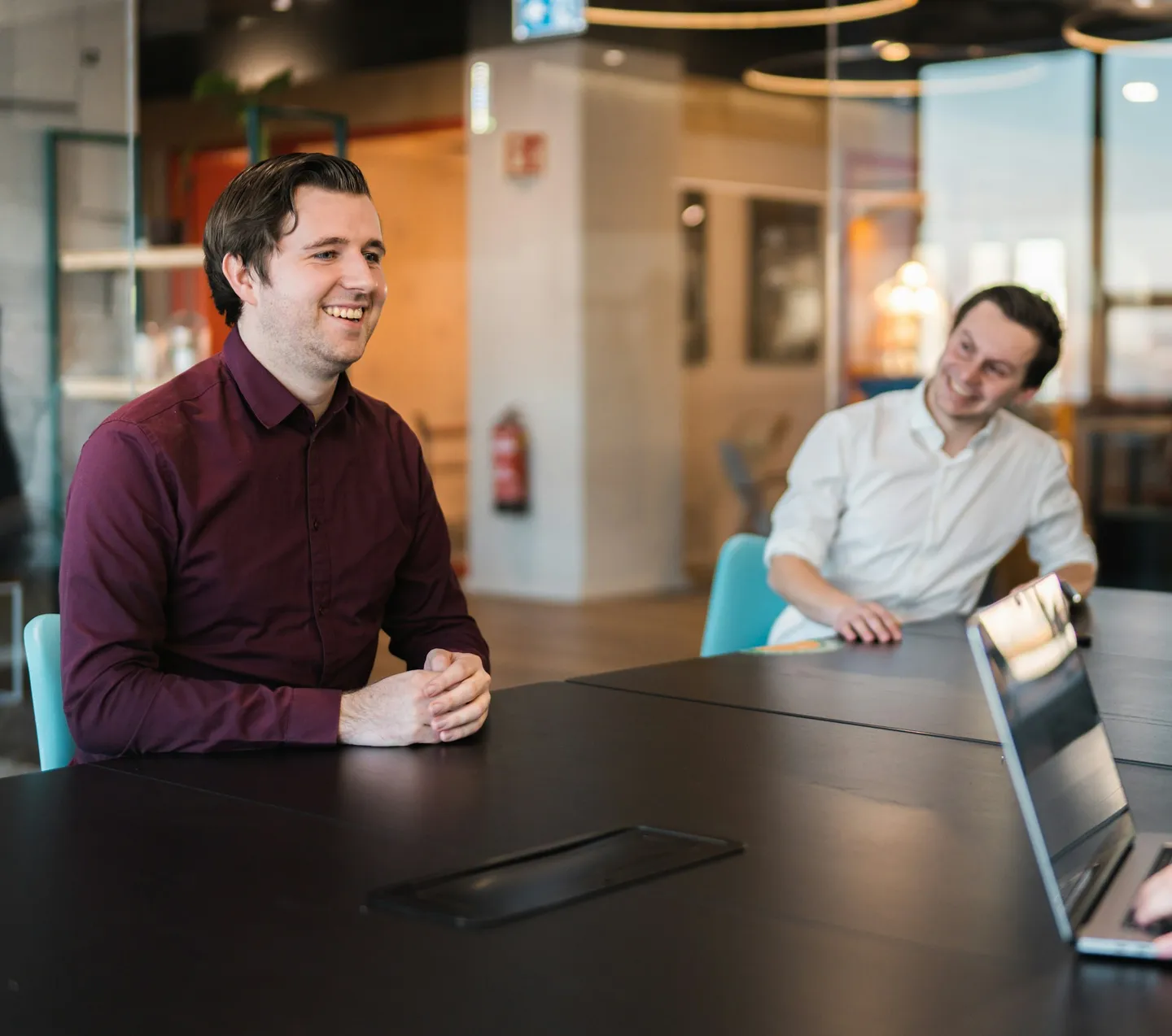 Twee lachende mannen aan een vergadertafel met een laptop